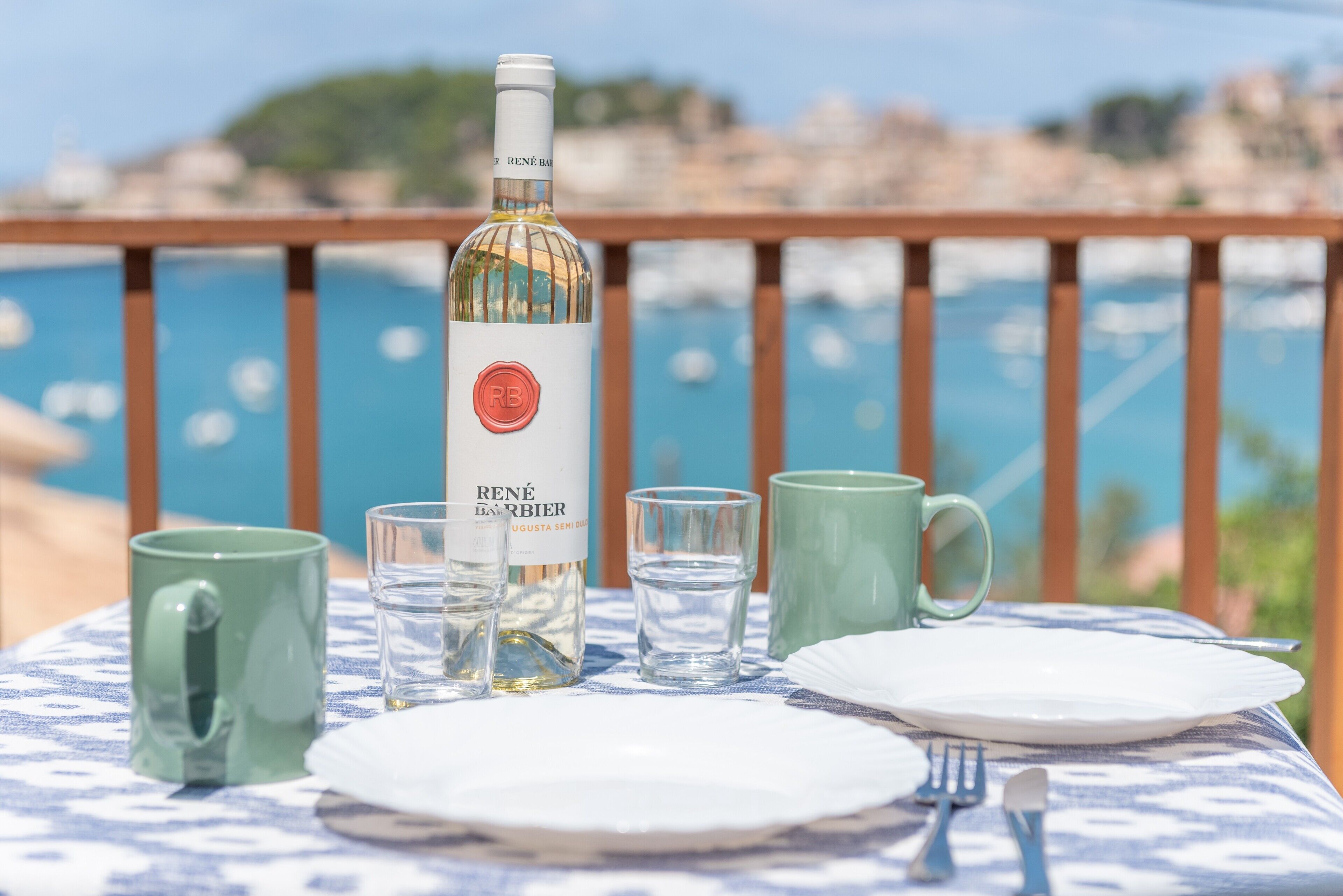 Una mesa dispuesta con una botella de vino, copas y una taza verde sobre un mantel azul y blanco, con vistas a una masa de agua.