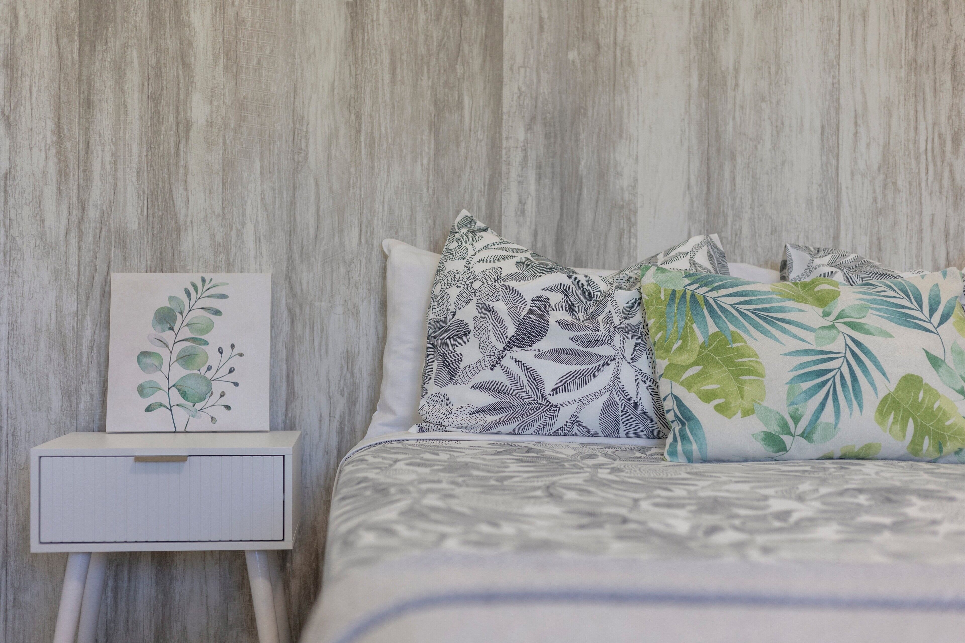 Uma cama com almofadas de padrão floral, uma mesa de cabeceira com gaveta e um quadro pendurado na parede.