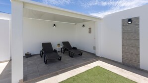 Een overdekt terras met twee loungestoelen, een kleine tafel met een plant en een betegelde vloer.