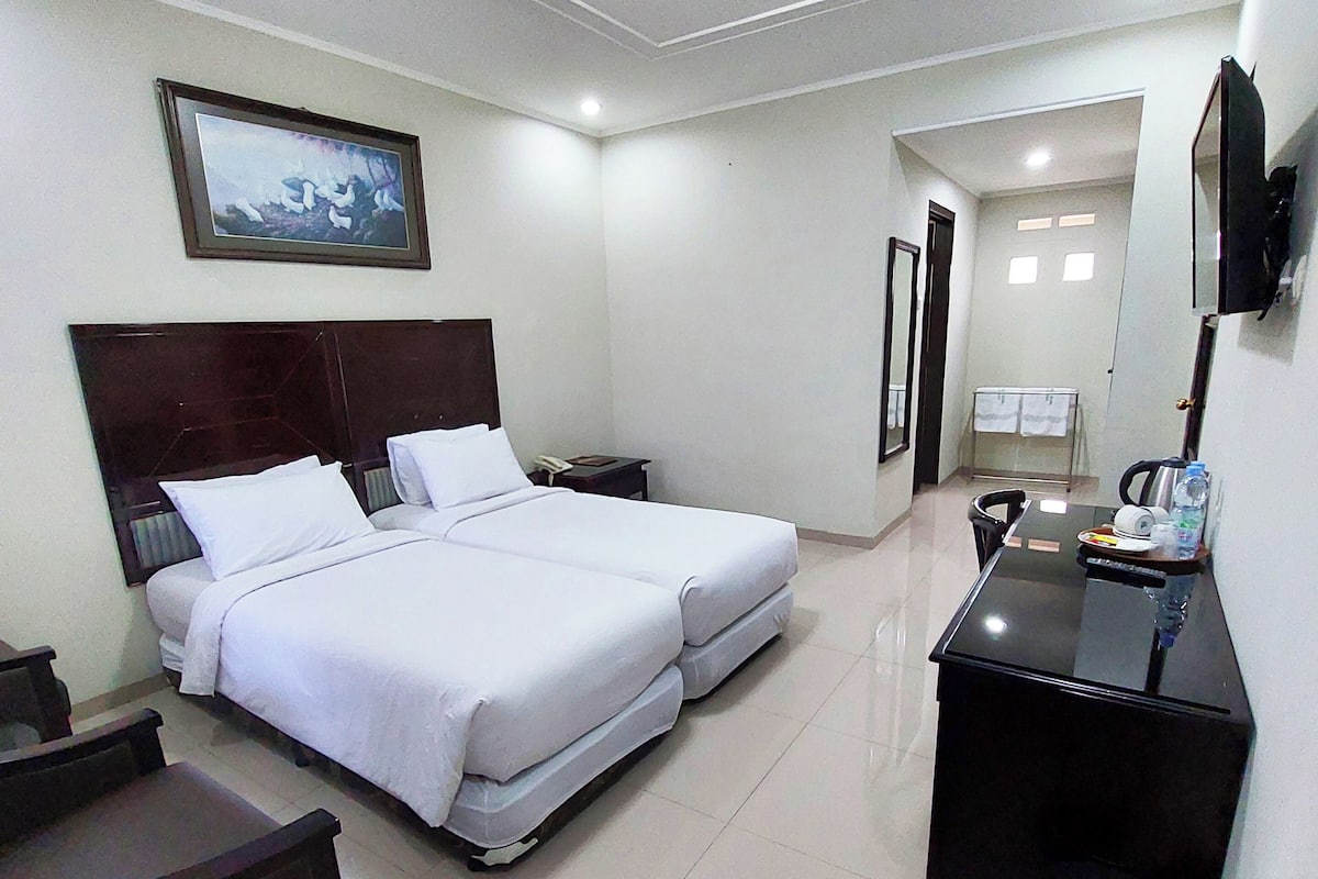 A hotel room with a bed, bedside tables, a desk, a trash bin, and a TV mounted on the wall.