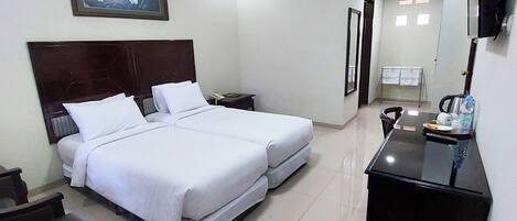 A hotel room with a bed, bedside tables, a desk, a trash bin, and a TV mounted on the wall.