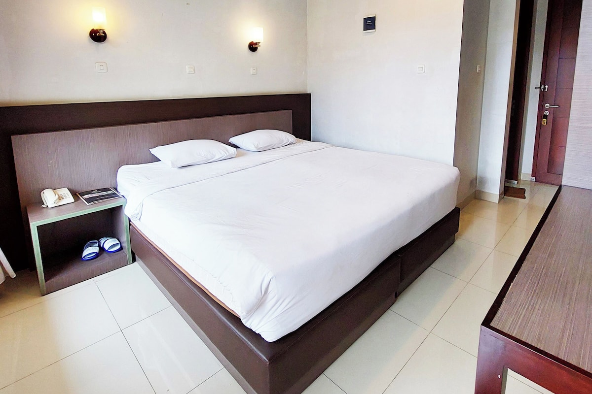 A hotel room with a large bed, a bedside table with a phone and a lamp, and a small shelf with shoes.