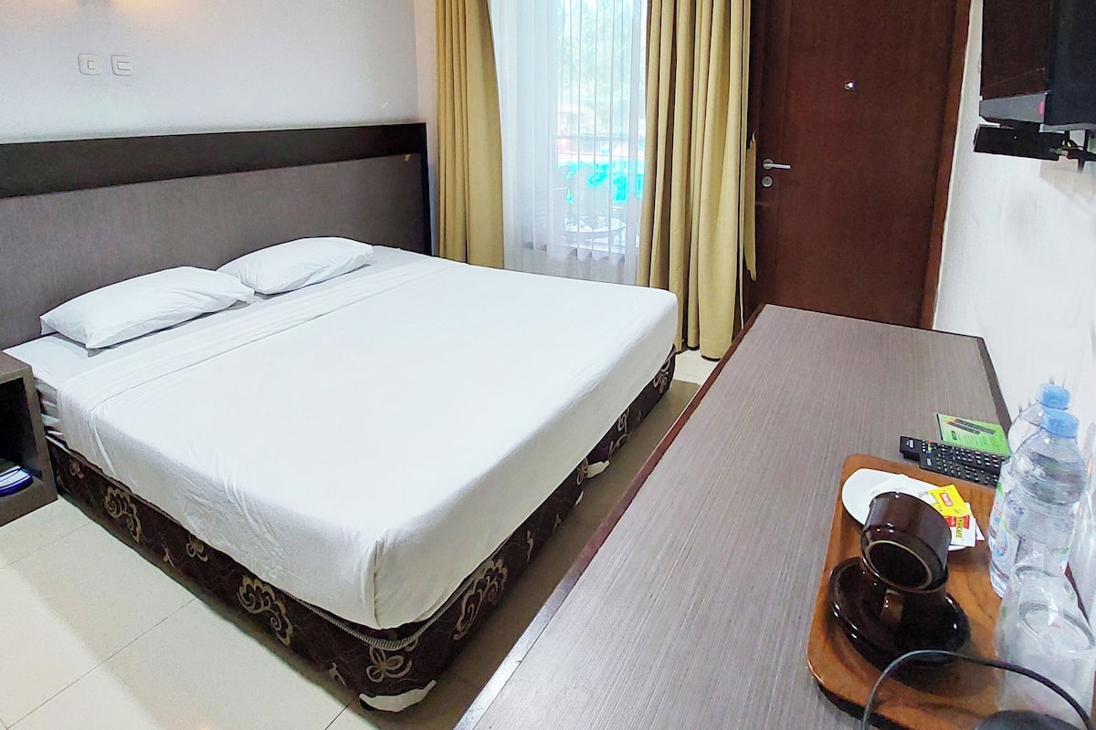 A hotel room with a bed, a desk with a computer, a chair, a TV, and a window with curtains.