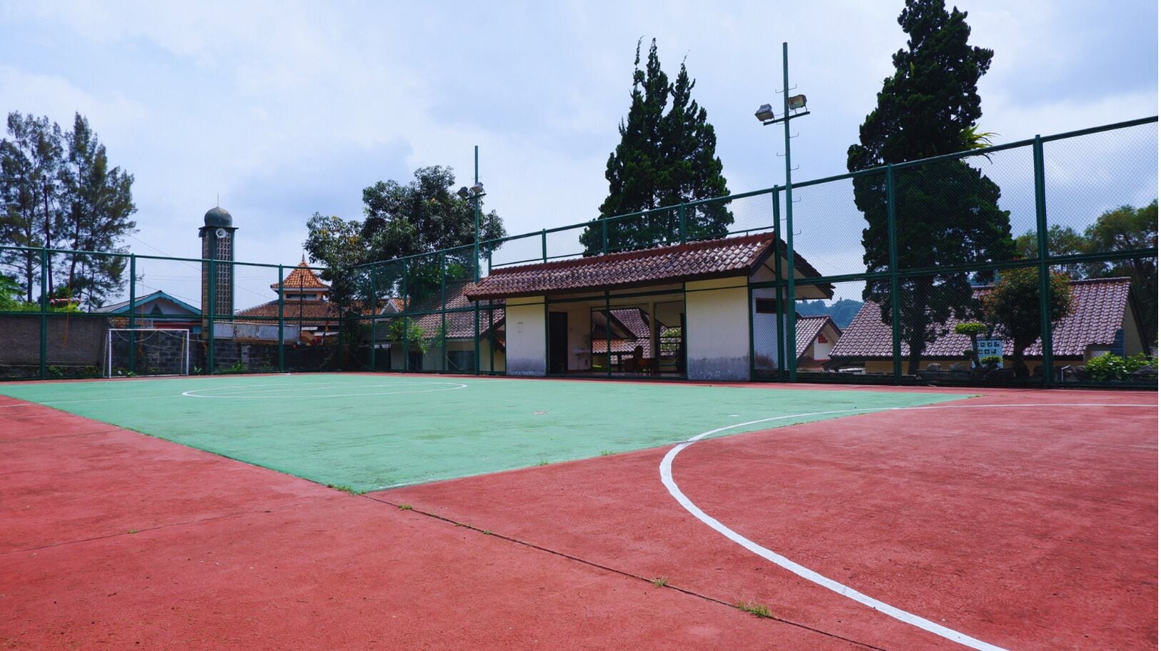 Ein Sportplatz im Freien mit grüner und roter Oberfläche, umgeben von einem Zaun und Bäumen.