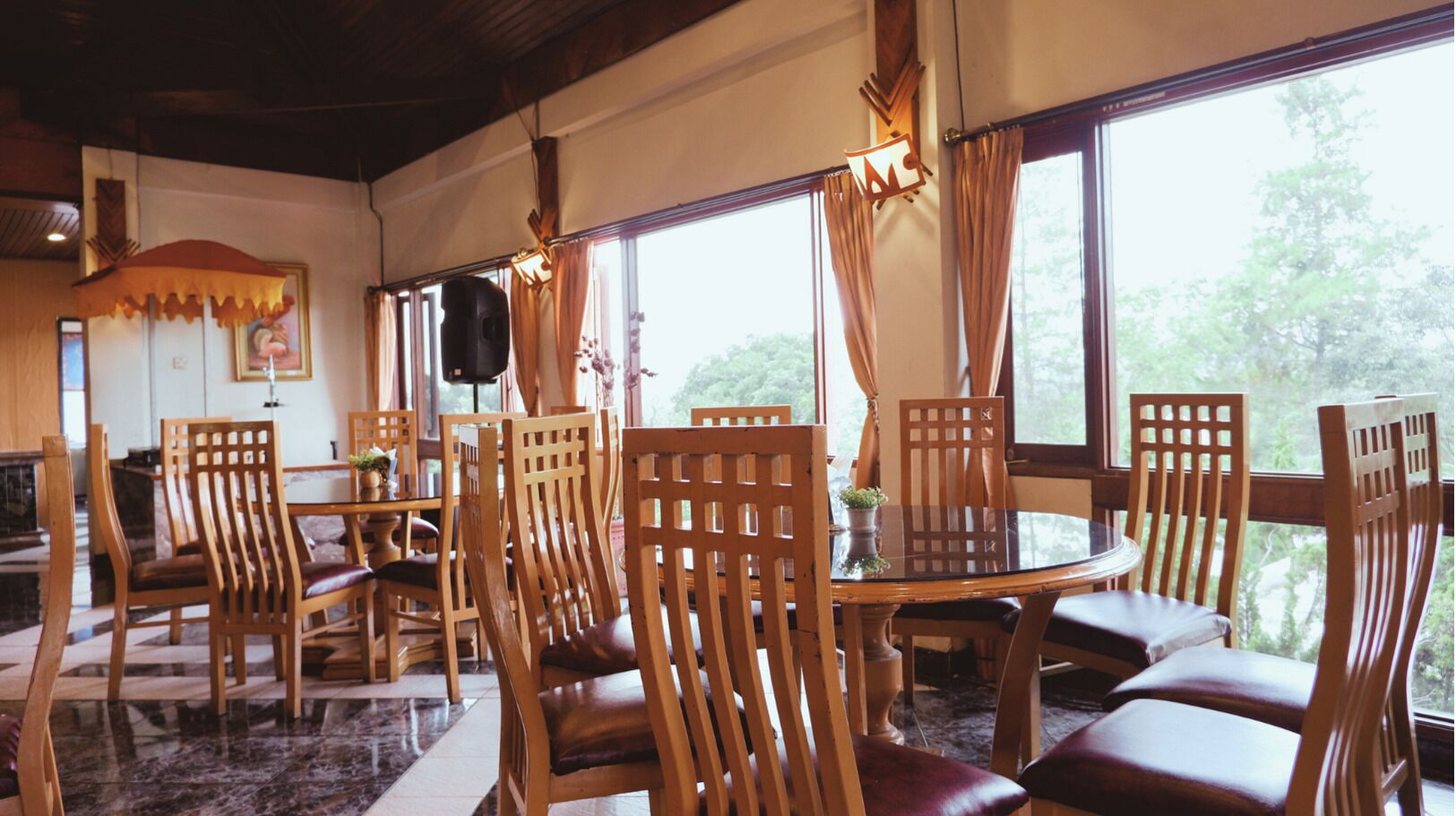 A dining area with wooden chairs and a round table, large windows with curtains, and a wall-mounted speaker.