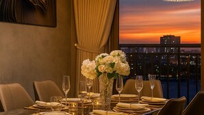 Un coin repas avec une table dressée pour quatre personnes, un vase rempli de fleurs blanches et une vue sur la ville au coucher du soleil.