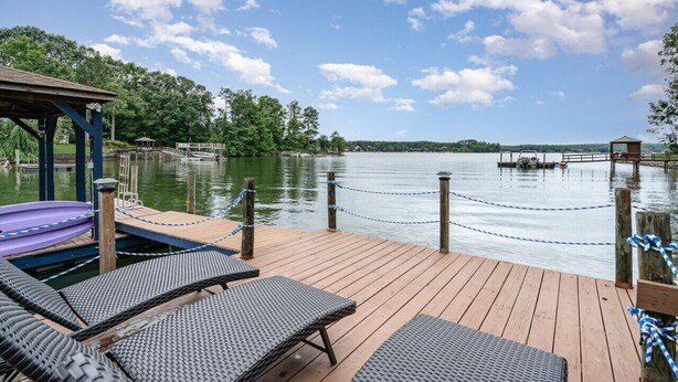 Un muelle de madera con sillones de descanso y un kayak, con vistas a un lago tranquilo con una embarcación amarrada y una estructura pequeña a lo lejos.