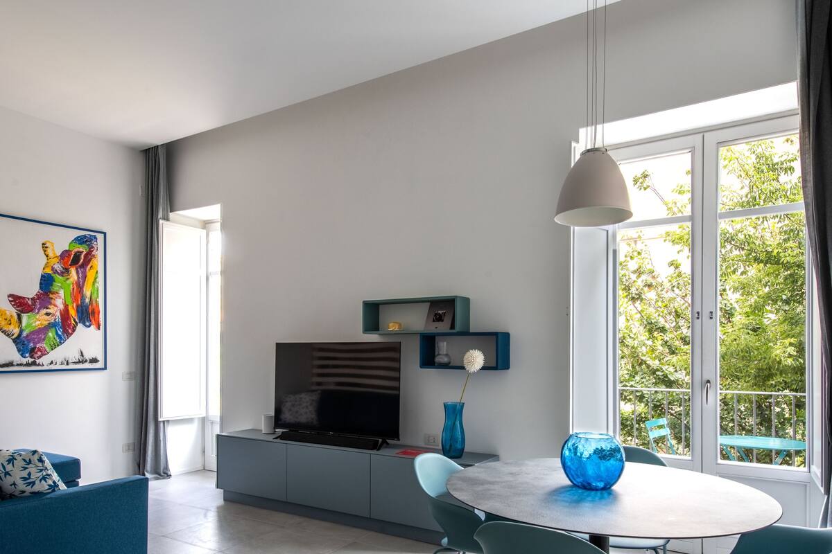 Uma sala de estar moderna com um sofá, uma mesa com cadeiras, uma televisão e uma janela grande com cortinas.