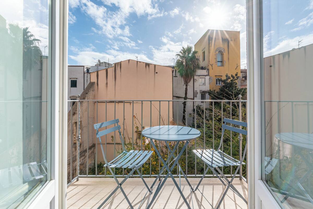 Varanda com mesa de vidro e duas cadeiras, com vista para área residencial com edifícios e uma palmeira.
