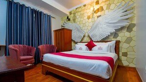 A hotel room with a large bed, two armchairs, a wooden table, and a wall with a winged angel decoration. - Hotel Parv (Jaipur)
