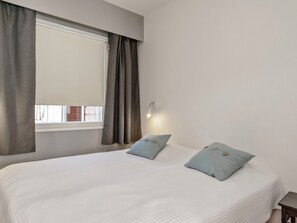 A bedroom with a large bed, two pillows, a nightstand, a window with curtains, and a wall-mounted light.