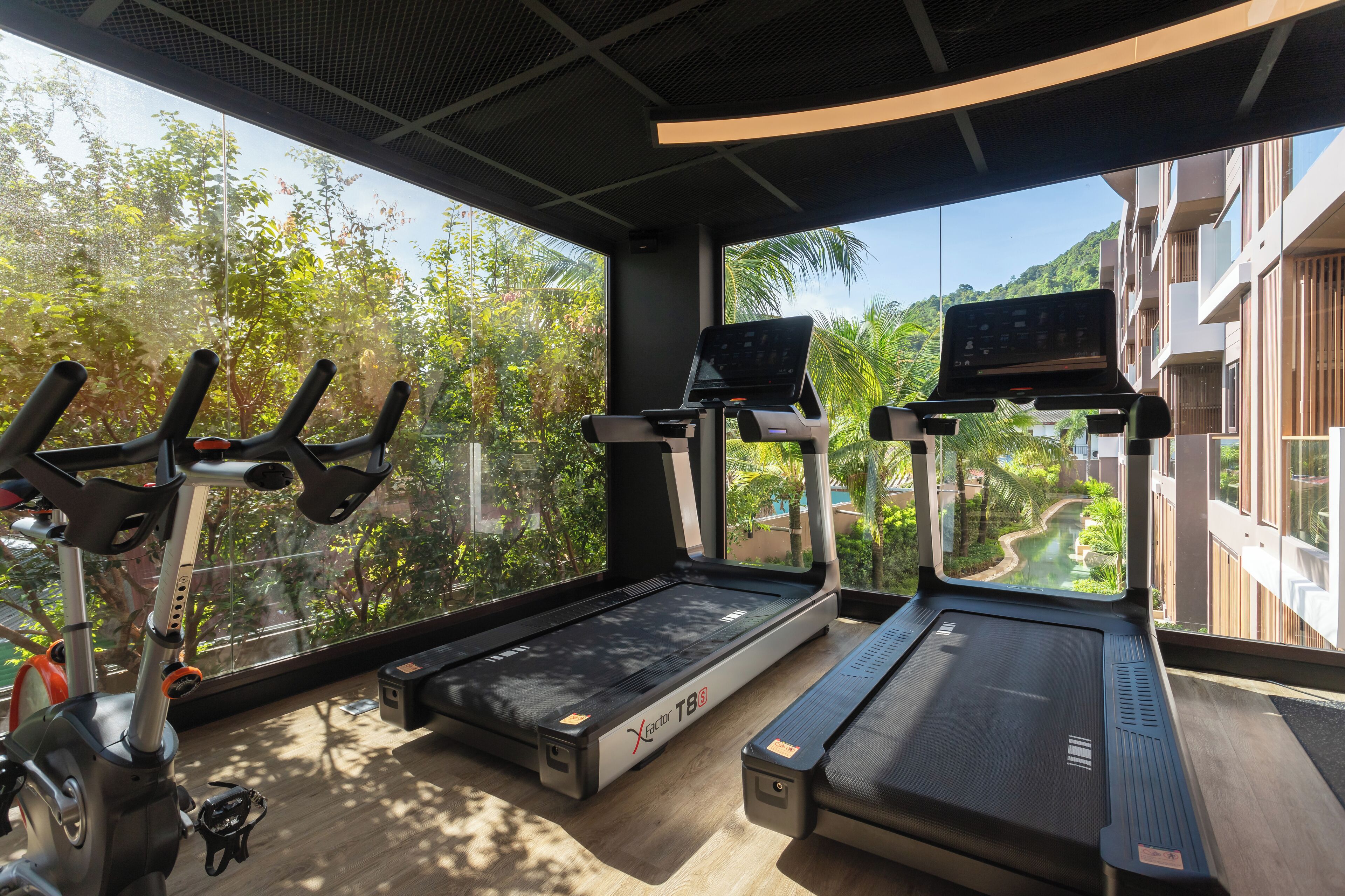 Une salle de sport équipée de tapis de course, de vélos d’appartement et d’un rameur, avec de grandes fenêtres offrant une vue sur la verdure et les bâtiments environnants.