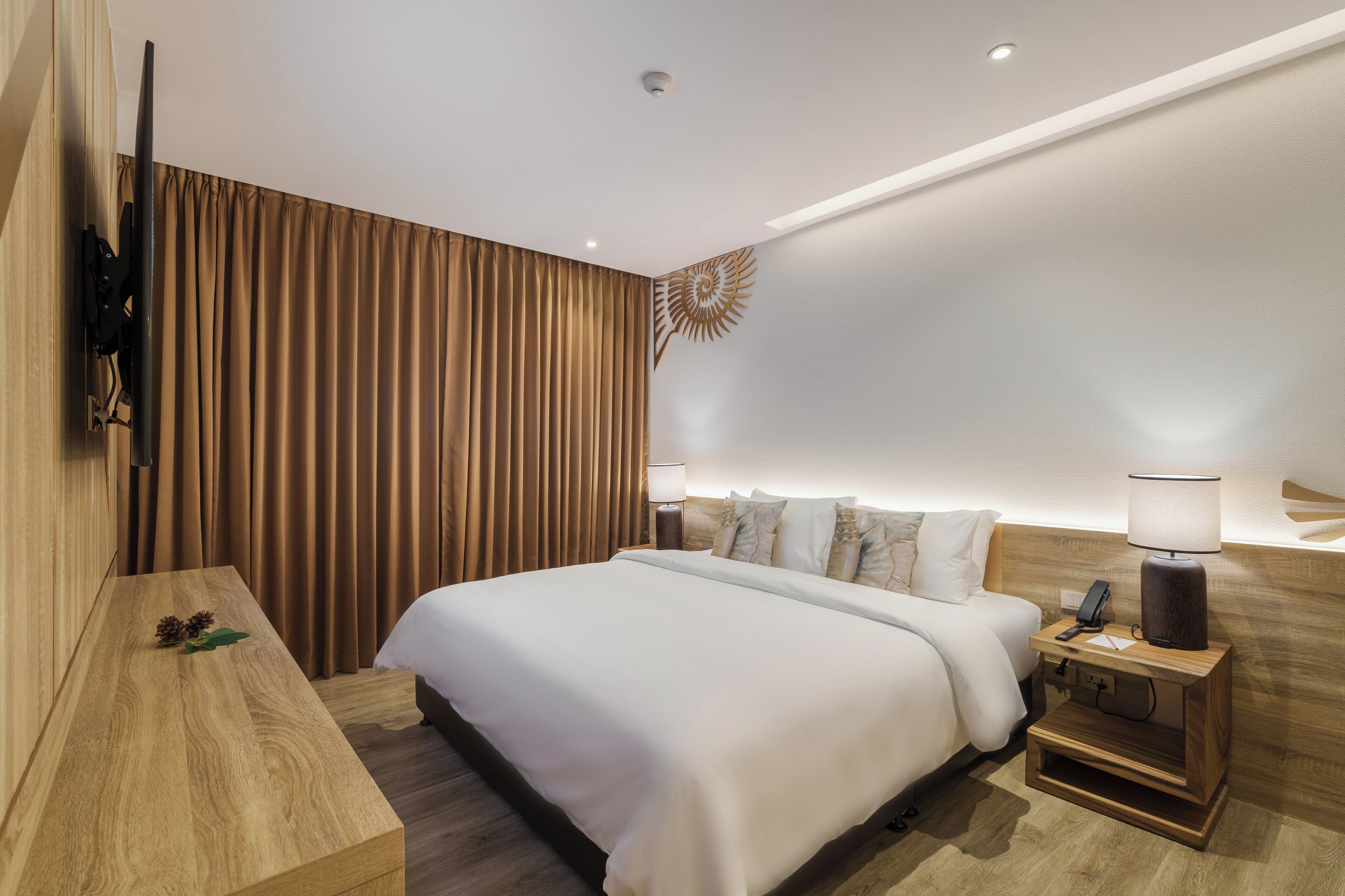 A modern hotel room with a large bed, wooden headboard, a nightstand with a lamp, and a wall-mounted TV.