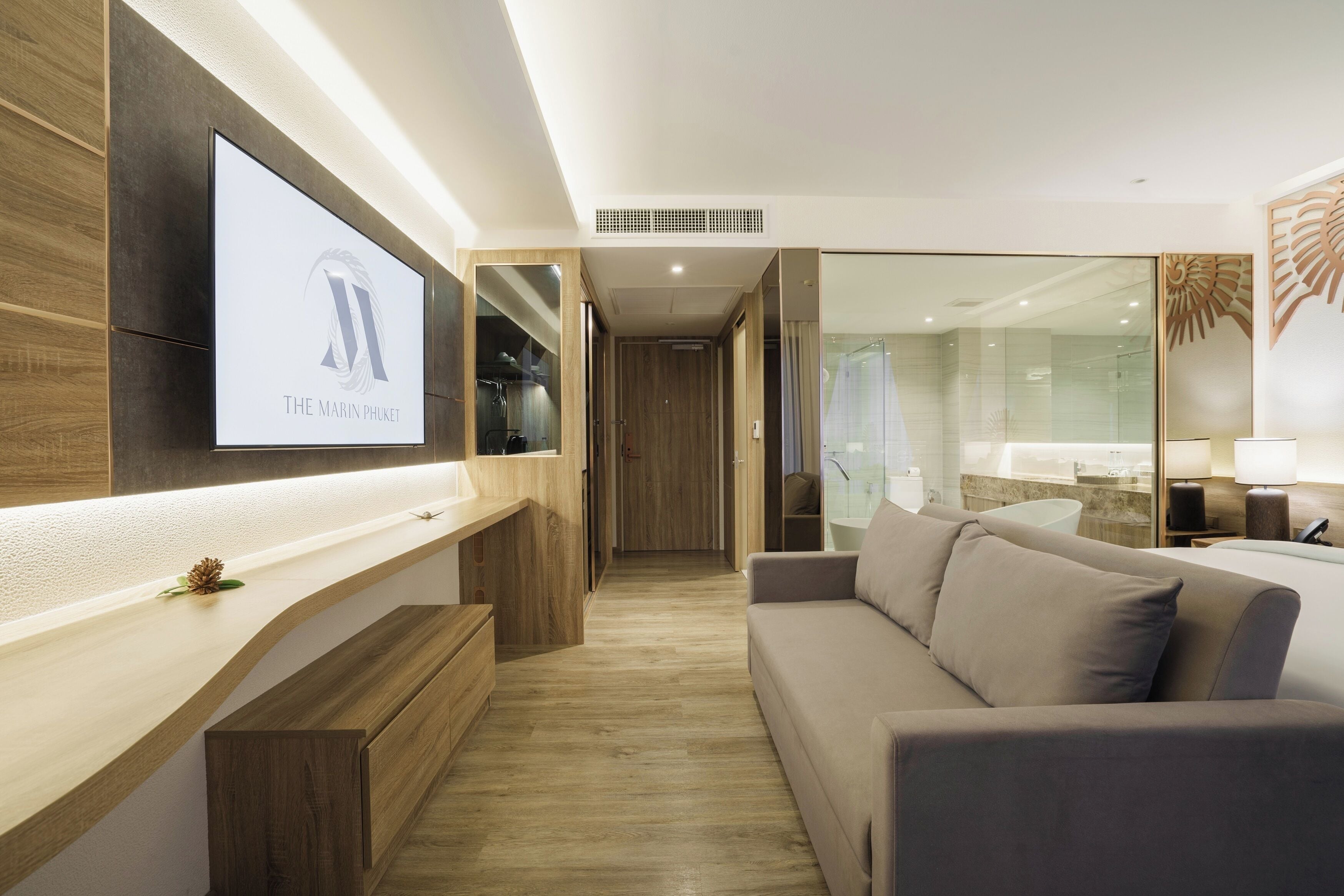 Una habitación de hotel moderna con piso de madera, un sofá, un televisor montado en la pared y una zona de ducha de vidrio.