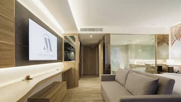 A modern hotel room with a wooden floor, a sofa, a TV mounted on the wall, and a glass shower area.
