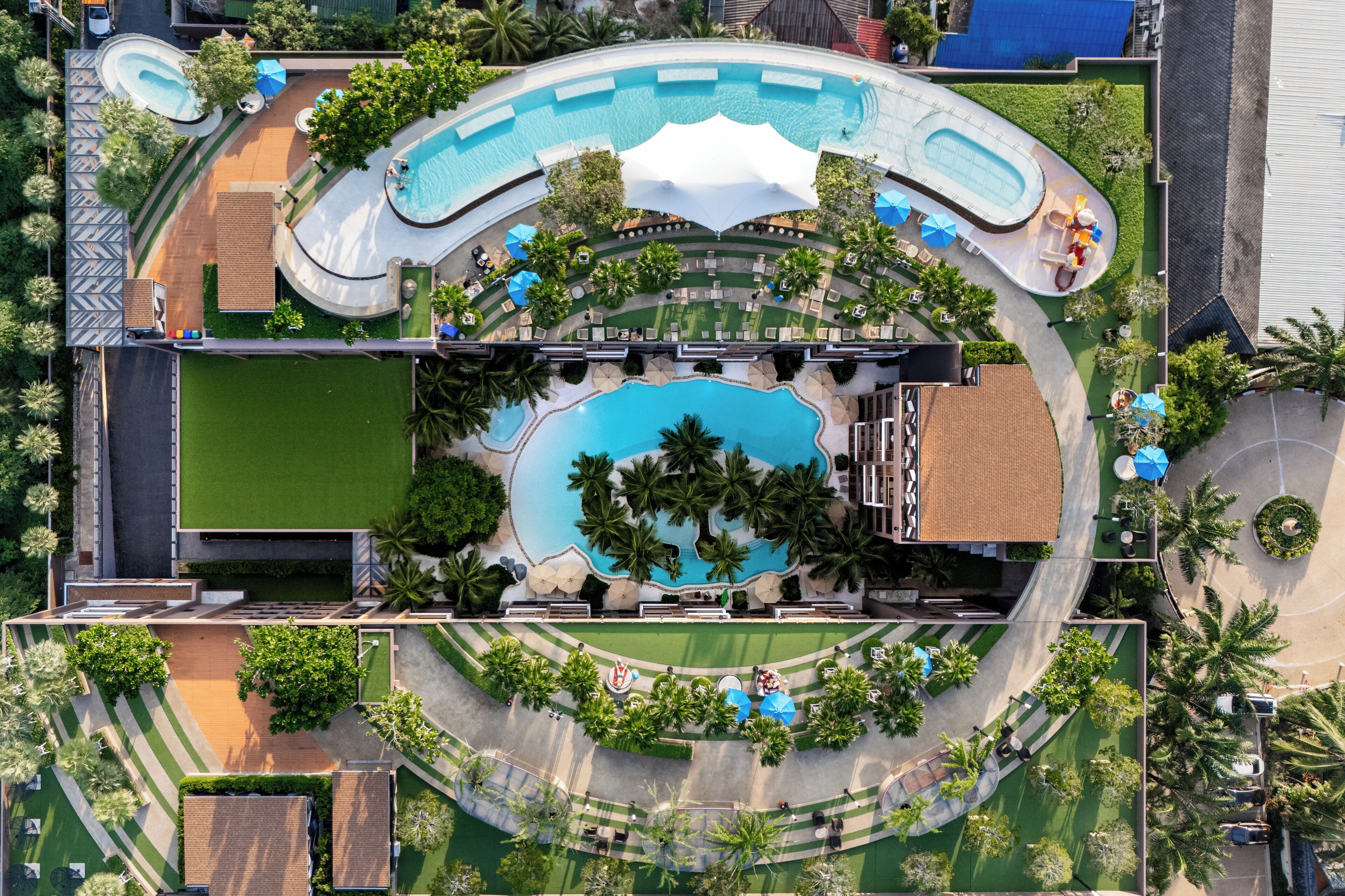 An aerial view of a resort with a large central pool, multiple smaller pools, and a variety of outdoor seating areas surrounded by palm trees and greenery.