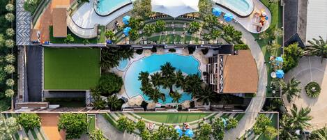 An aerial view of a resort with a large central pool, multiple smaller pools, and a variety of outdoor seating areas surrounded by palm trees and greenery.