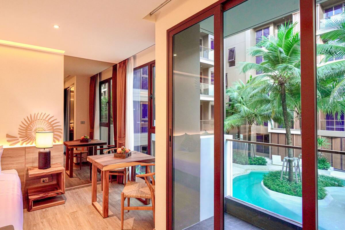 A hotel room with a bed, a desk, a chair, a lamp, and a large window overlooking a pool and palm trees.