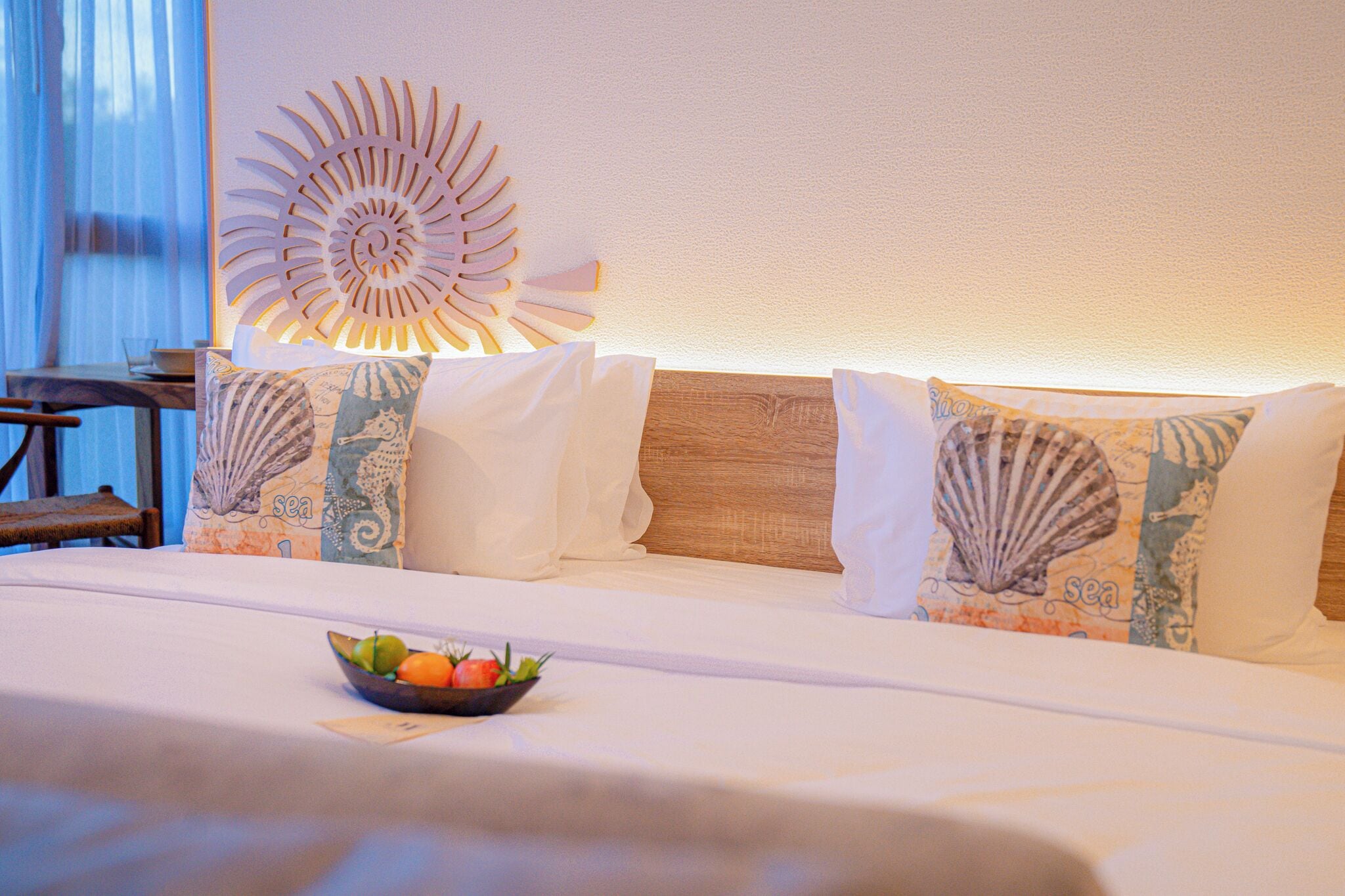 A hotel room with a bed, pillows, a wall decoration, and a bowl of fruit on the bed.