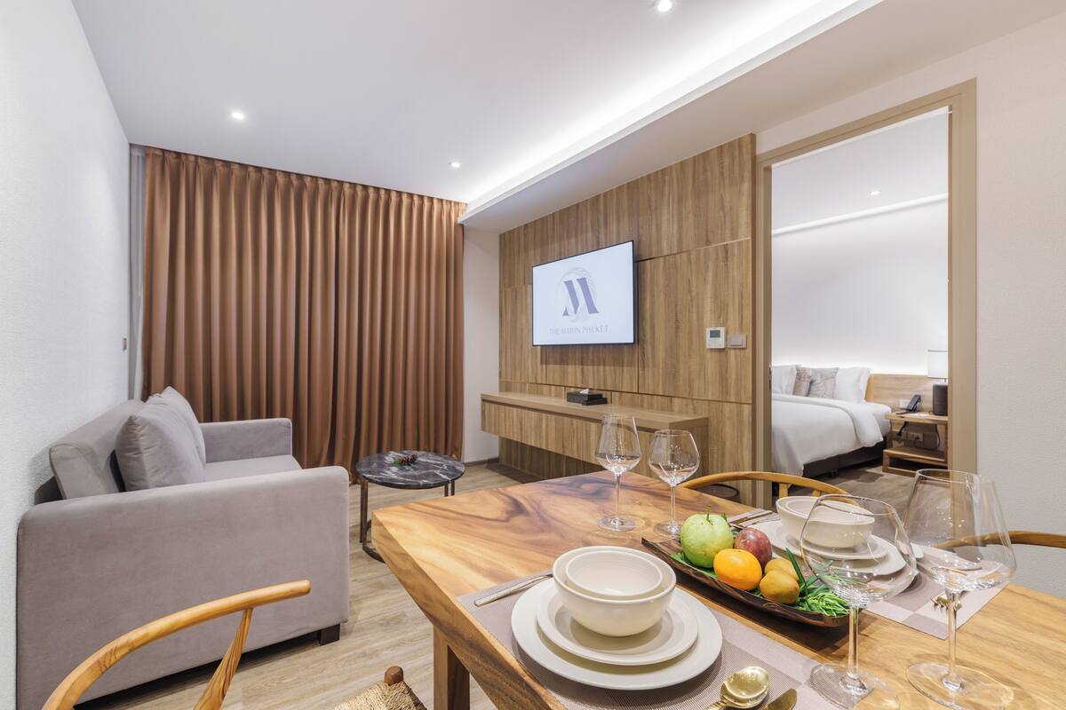 A modern hotel room with a wooden accent wall, a flat-screen TV, a dining area with a fruit tray, and a sofa.