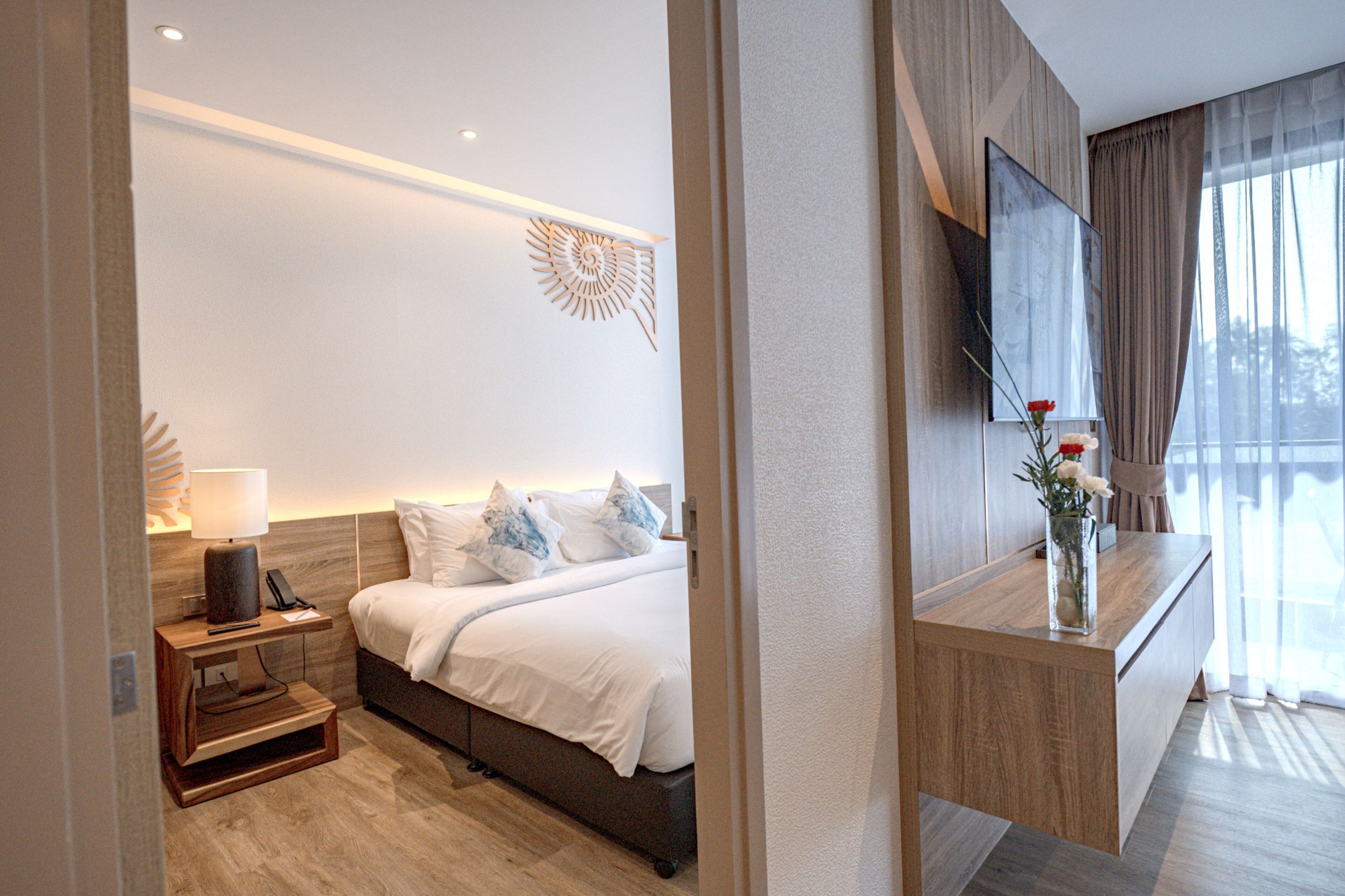 Quarto de hotel moderno com cama, mesa de cabeceira, luminária e televisão sobre um console de madeira.