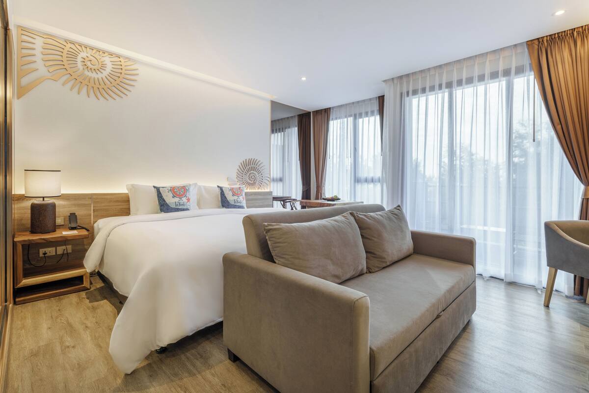 A modern hotel room with a large bed, a sofa, a nightstand, and a lamp.