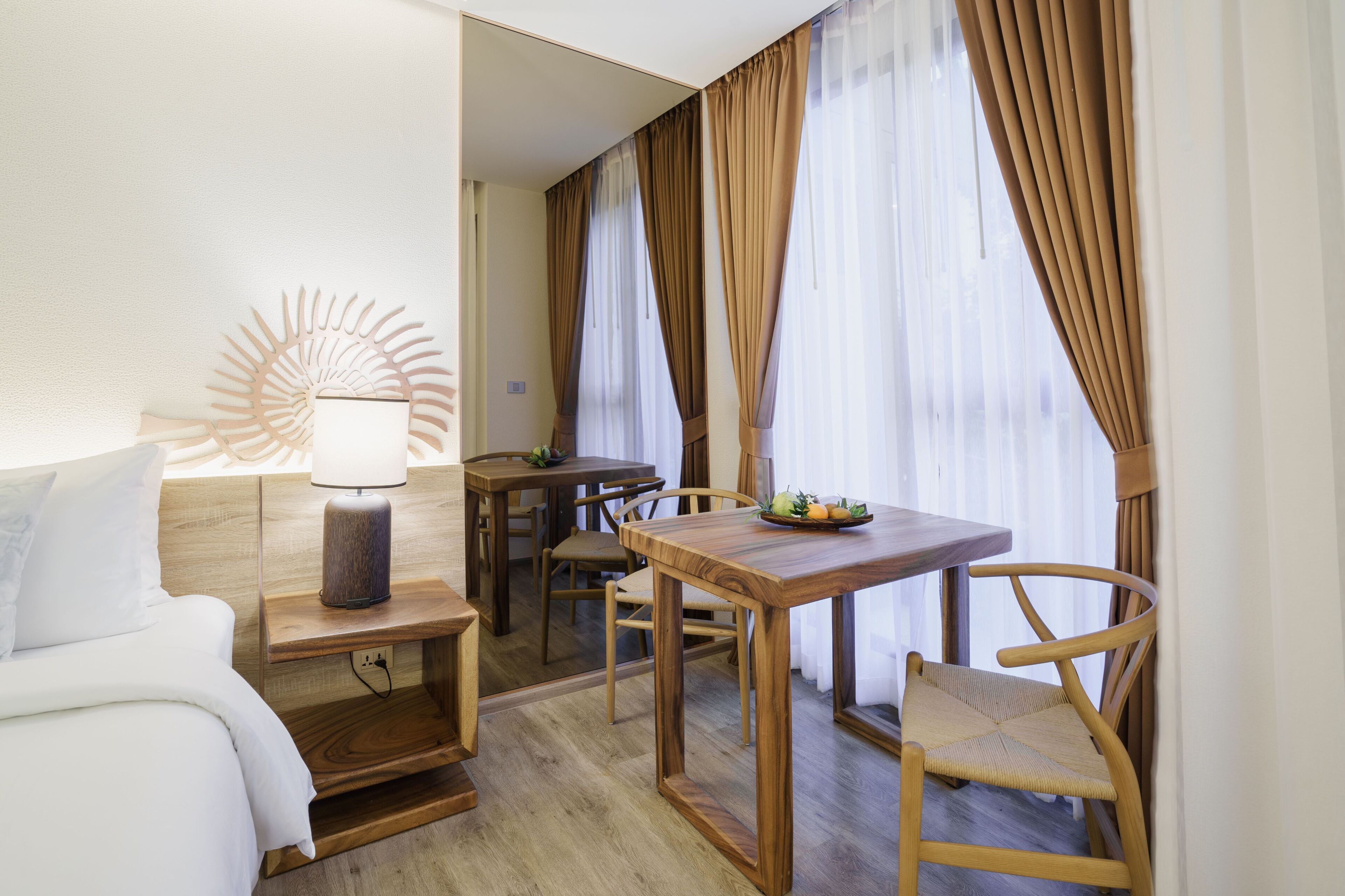 A hotel room with a bed, bedside table with a lamp, a wooden dining table with chairs, and a view through sheer curtains.