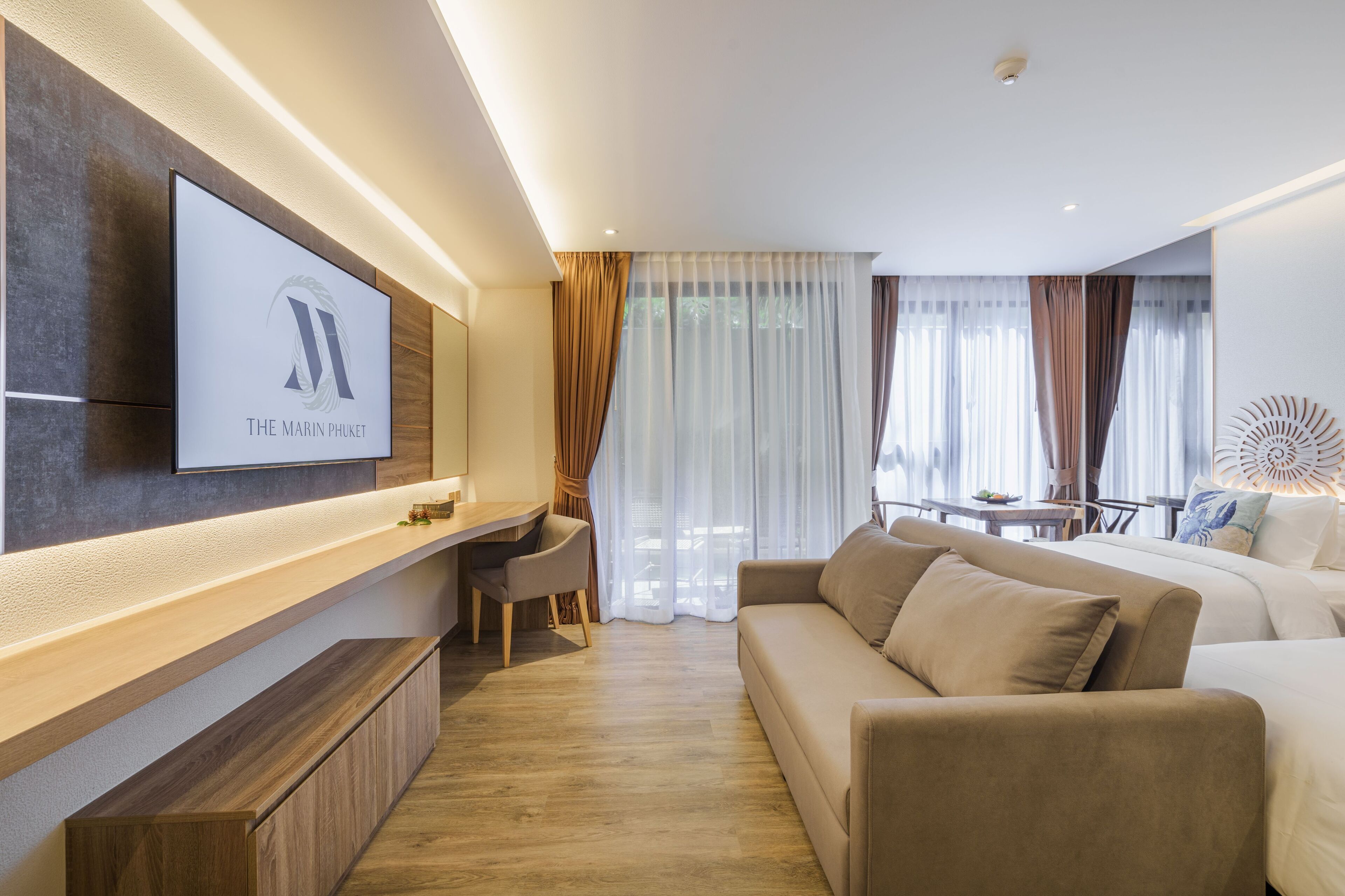 Moderni hotellihuone, jossa on seinälle asennettu taulutelevisio, sohva, työpöytä tuolilla sekä ruokailutila, jossa on pöytä ja tuolit.
