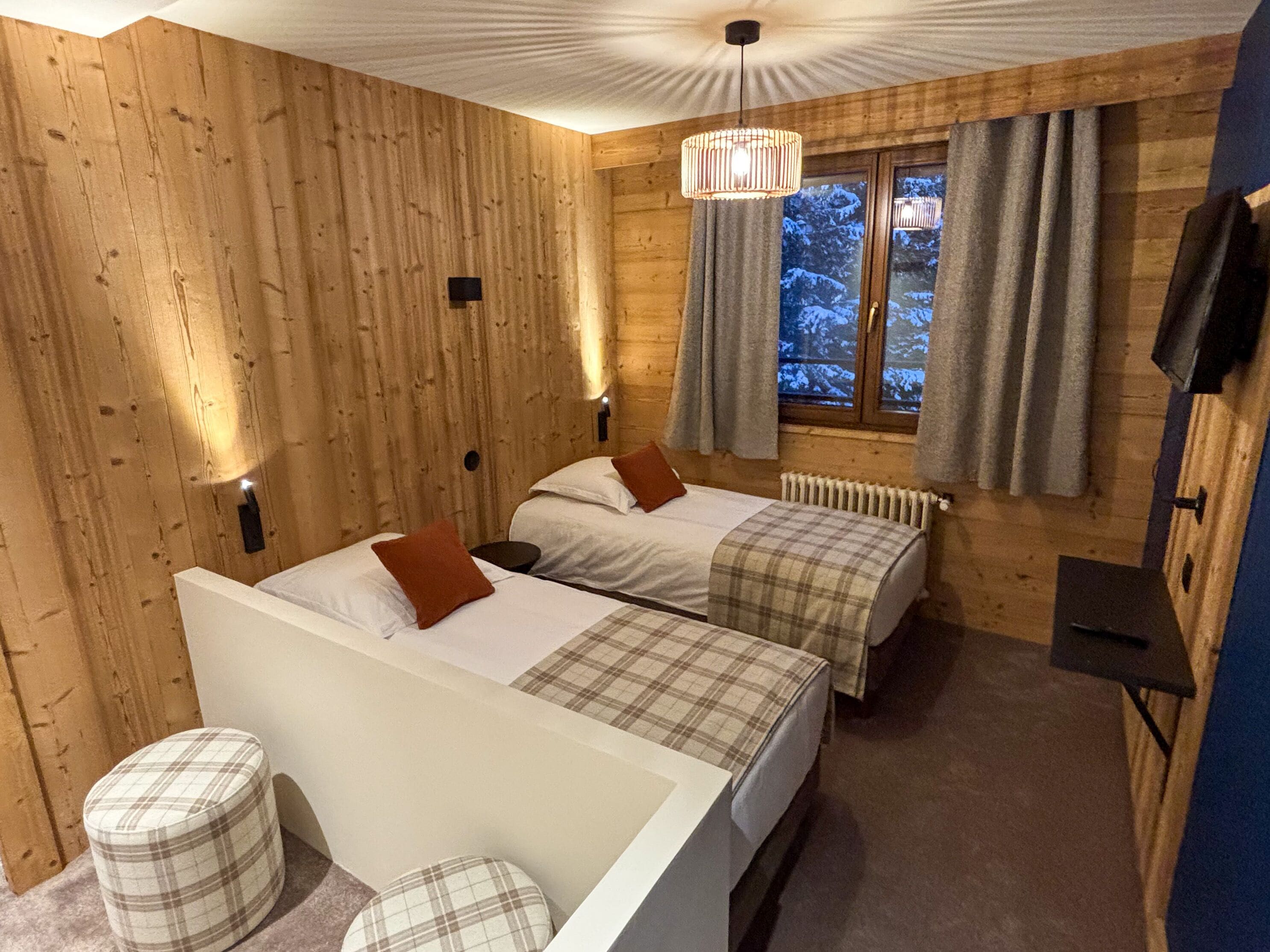 Ein Holzhüttenraum mit zwei Betten, einem kleinen Tisch und einem Fenster mit Vorhängen.