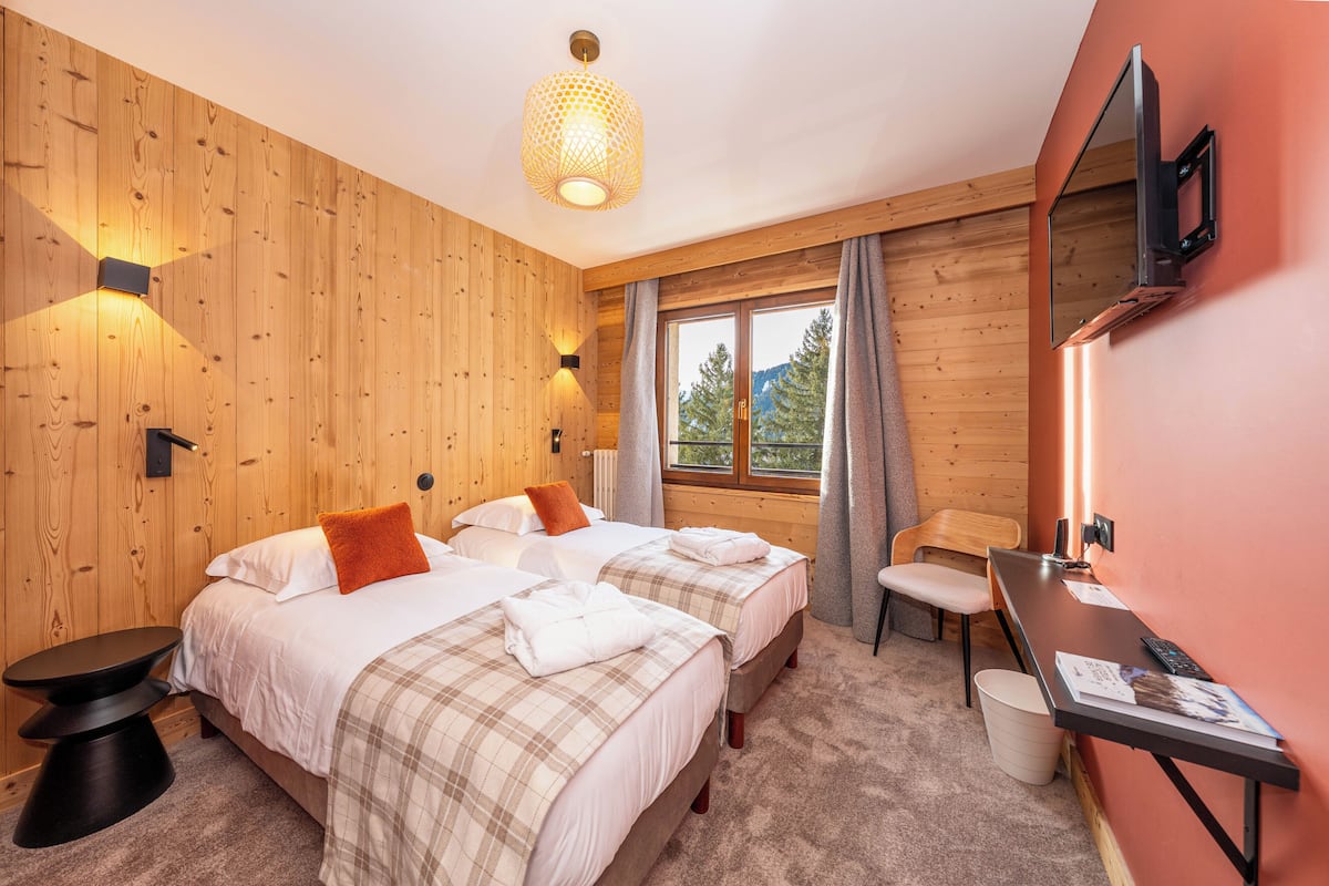 A room with two beds, a desk, a chair, a small table, and a window with a view of trees.