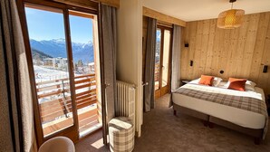 A bedroom with a bed, a chair, a radiator, and a view of mountains through large windows. - Casa Moho Saint-François-Longchamp 1450 (Saint-François-Longchamp)