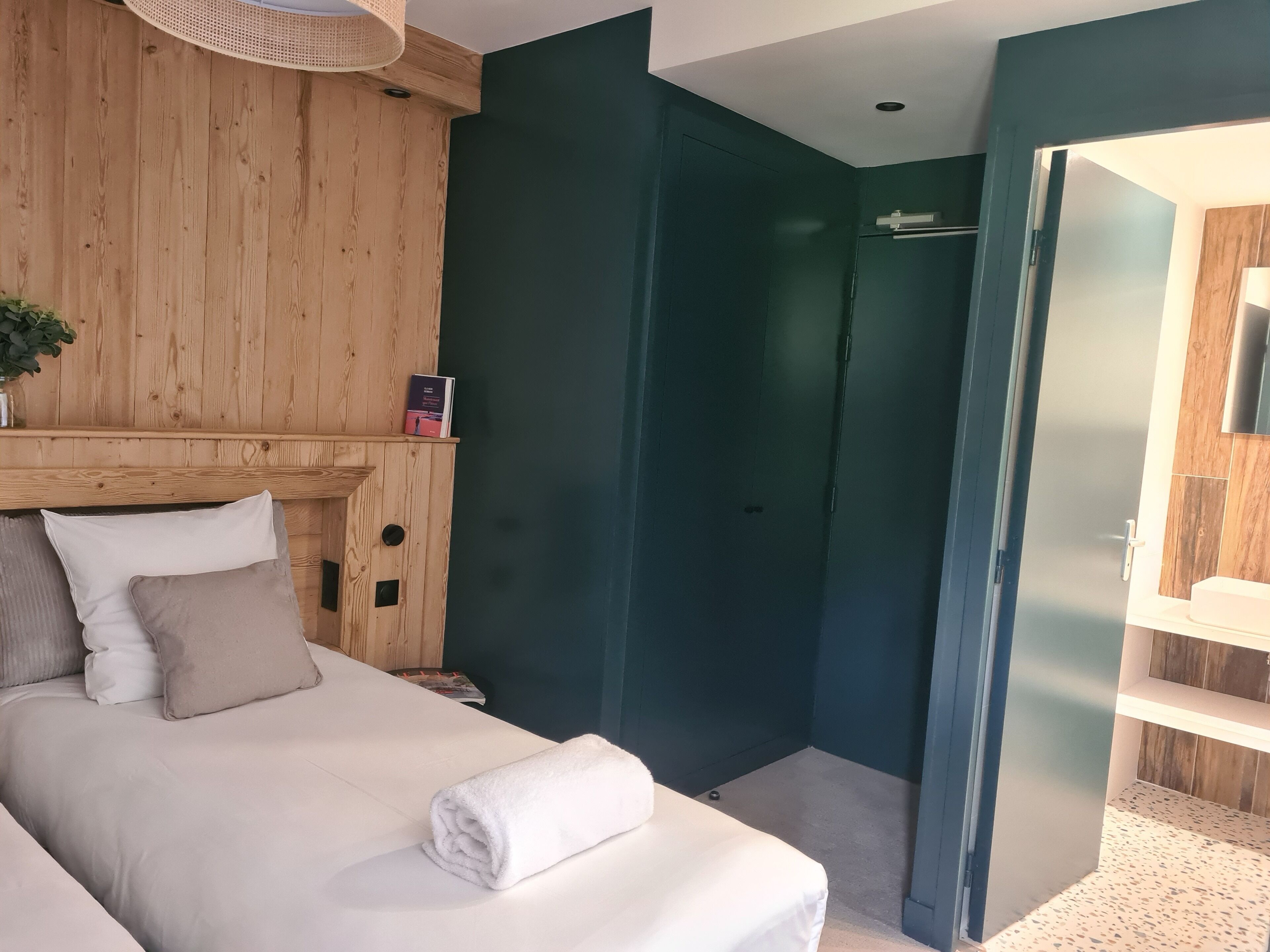 A bedroom with a bed, pillows, a bedside table, a wooden headboard, a dark green door, and a small shelf with a book.