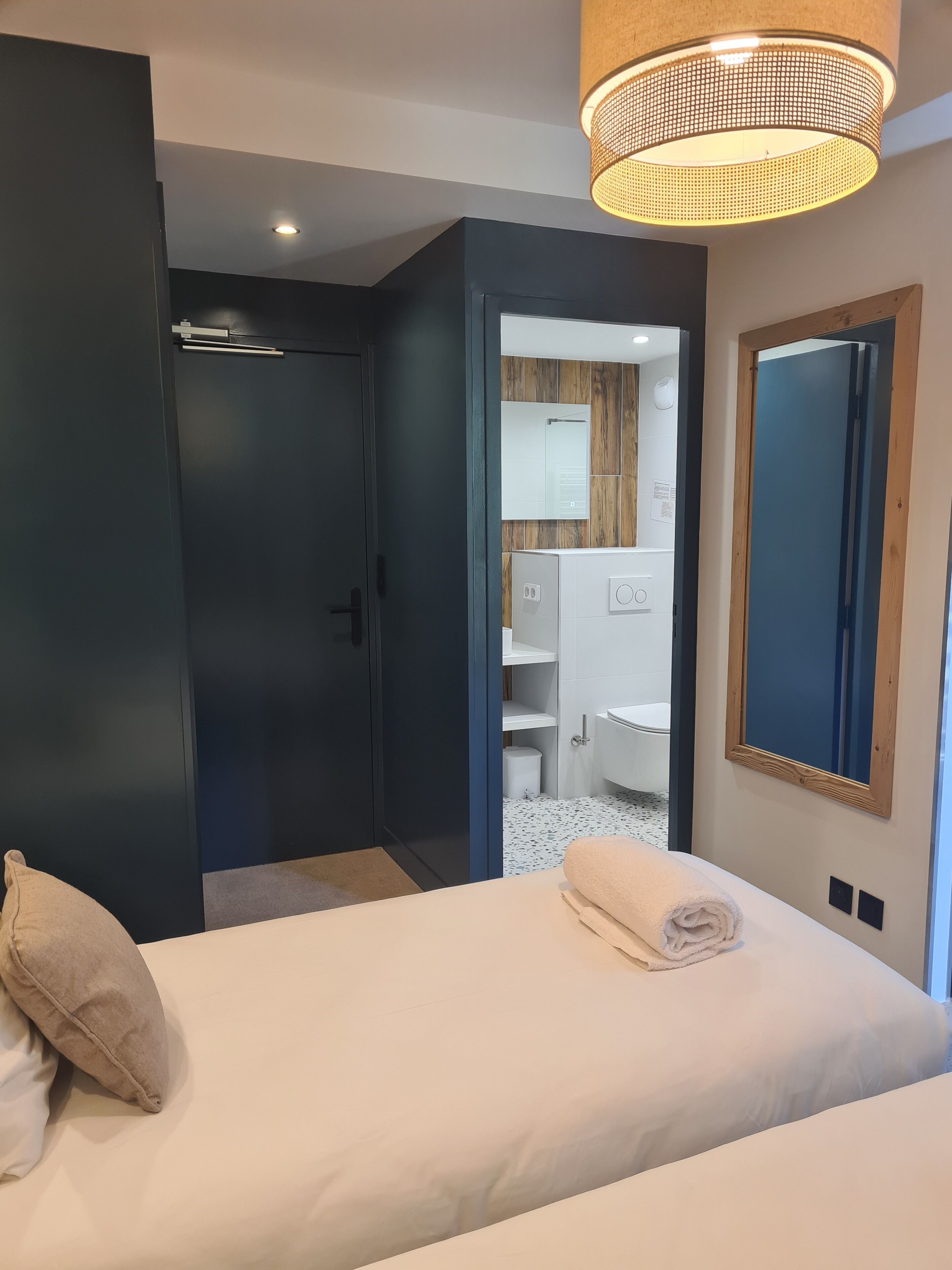 Una habitación de hotel con cama, un armario de color oscuro, un baño visible a través de una puerta abierta y un espejo grande en la pared.