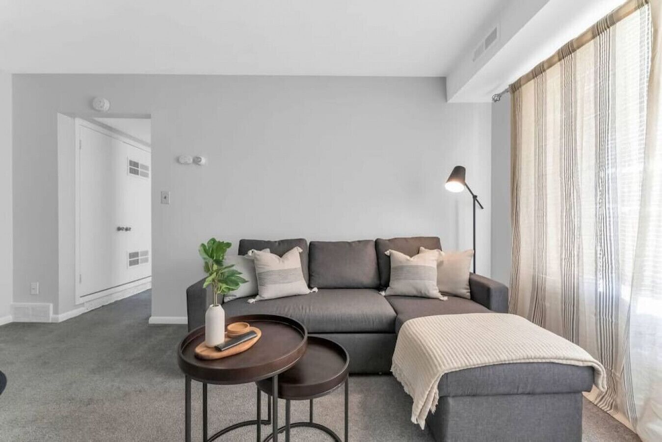 Una sala de estar moderna con un sofá gris, dos mesitas auxiliares, una lámpara de pie y un ventanal con cortinas translúcidas.