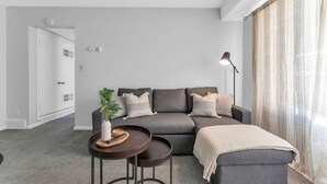 Un salon moderne avec un canapé gris, deux tables d’appoint, une lampe sur pied et une grande fenêtre ornée de rideaux légers.