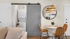 Un espacio habitable compacto con kitchenette, una zona de comedor con sillas de mimbre, una puerta corredera gris y un espejo redondo en la pared.