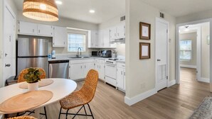 Una cocina moderna con gabinetes blancos, electrodomésticos de acero inoxidable, sillas de mimbre e iluminación colgante.