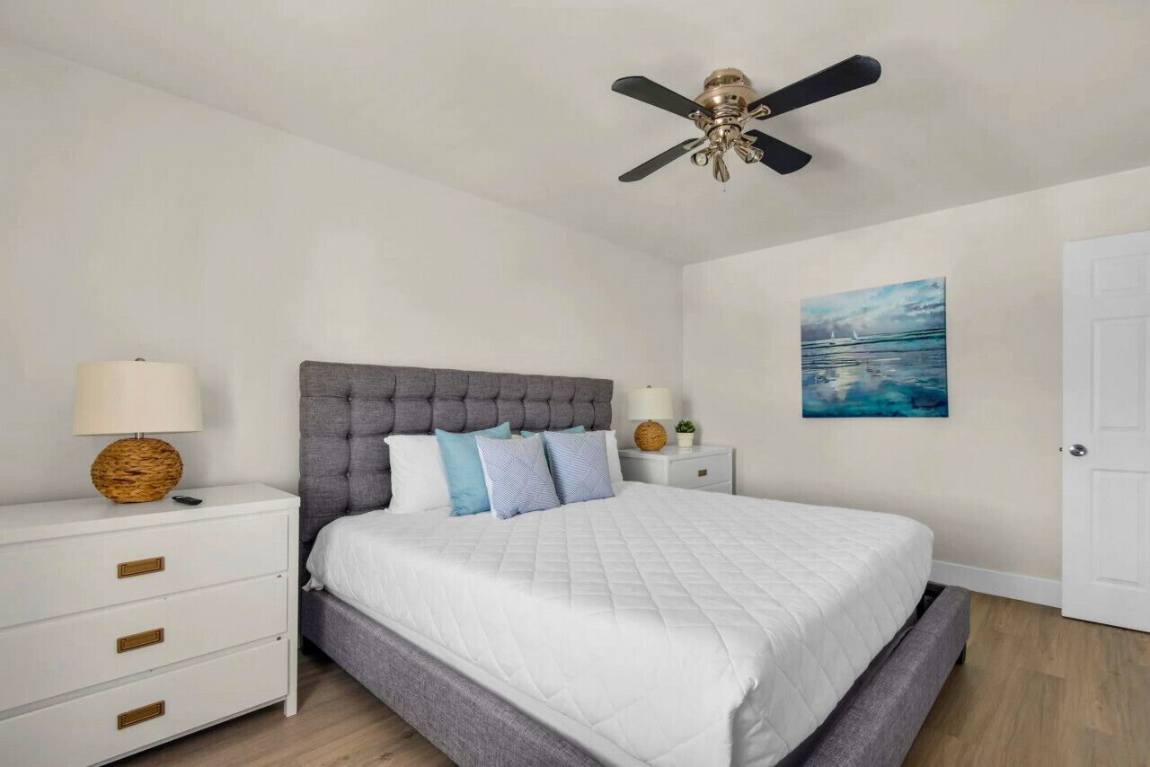 A bedroom with a tufted headboard, a bed with white bedding, a bedside table with a lamp, and a ceiling fan.