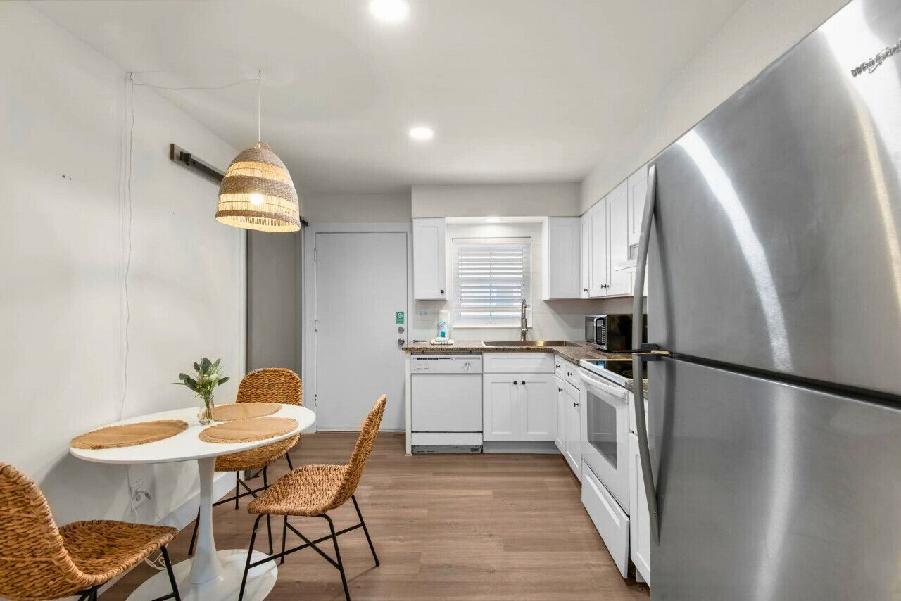 Cocina compacta con armarios blancos, refrigerador de acero inoxidable, microondas y comedor con sillas de mimbre y una mesa blanca.