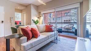 Un salón moderno con un sofá gris, cojines rojos, una mesita y un balcón con mobiliario exterior.