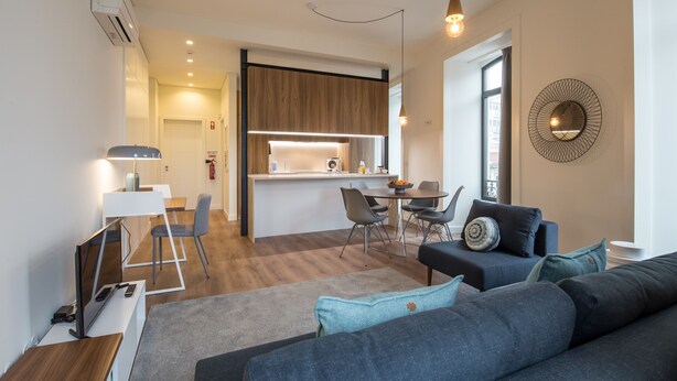 Een moderne woonkamer met een bank, een eethoek met stoelen, een keuken met een aanrecht en een groot raam.