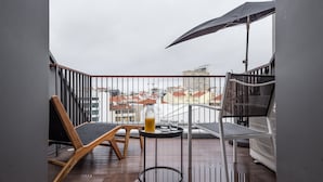 Ein Balkon mit einem Holzstuhl, einem kleinen runden Tisch, einem Glas und einer Flasche, sowie einem großen Sonnenschirm.