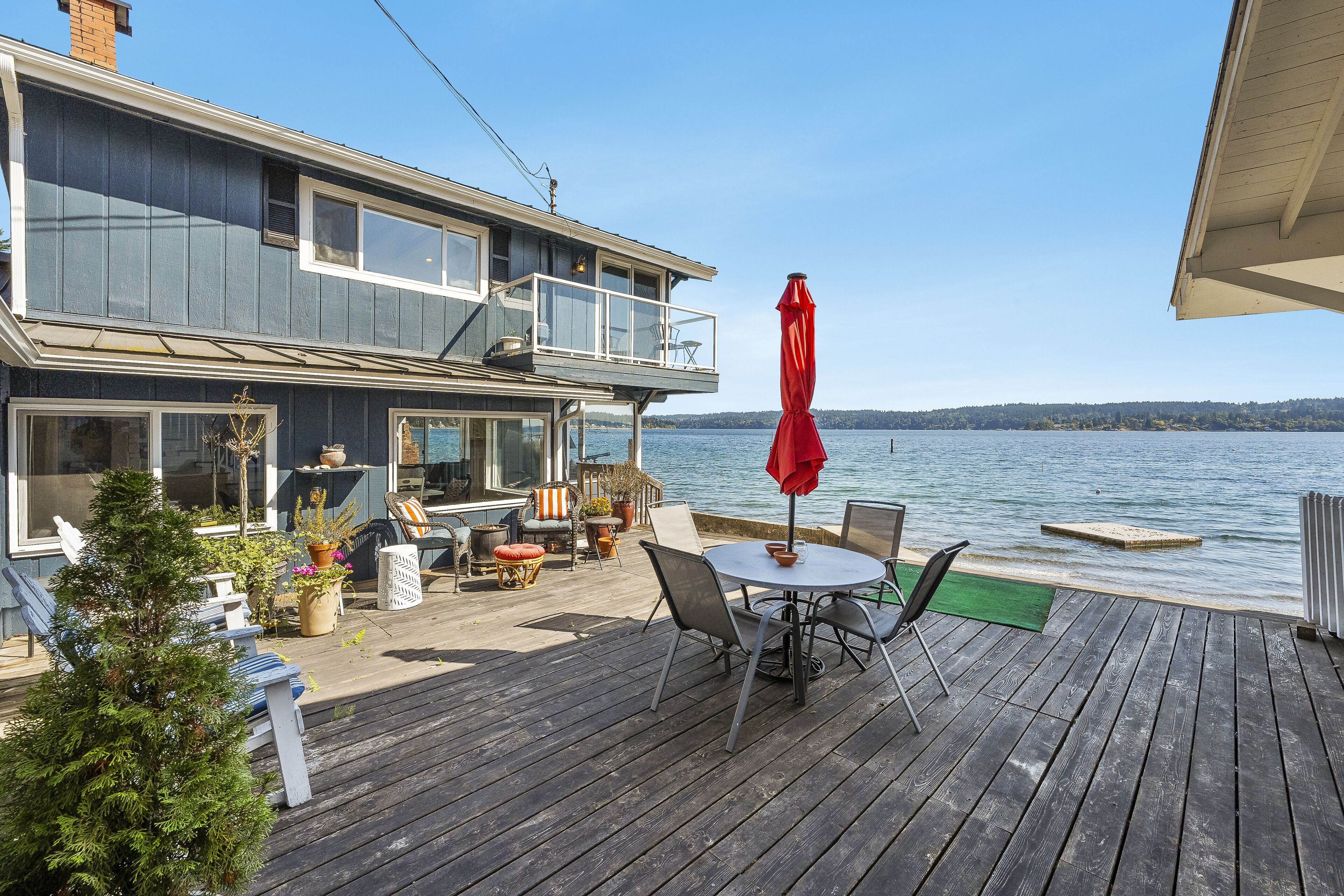 A two-story house with a deck overlooking a lake, featuring outdoor seating and a red umbrella.
