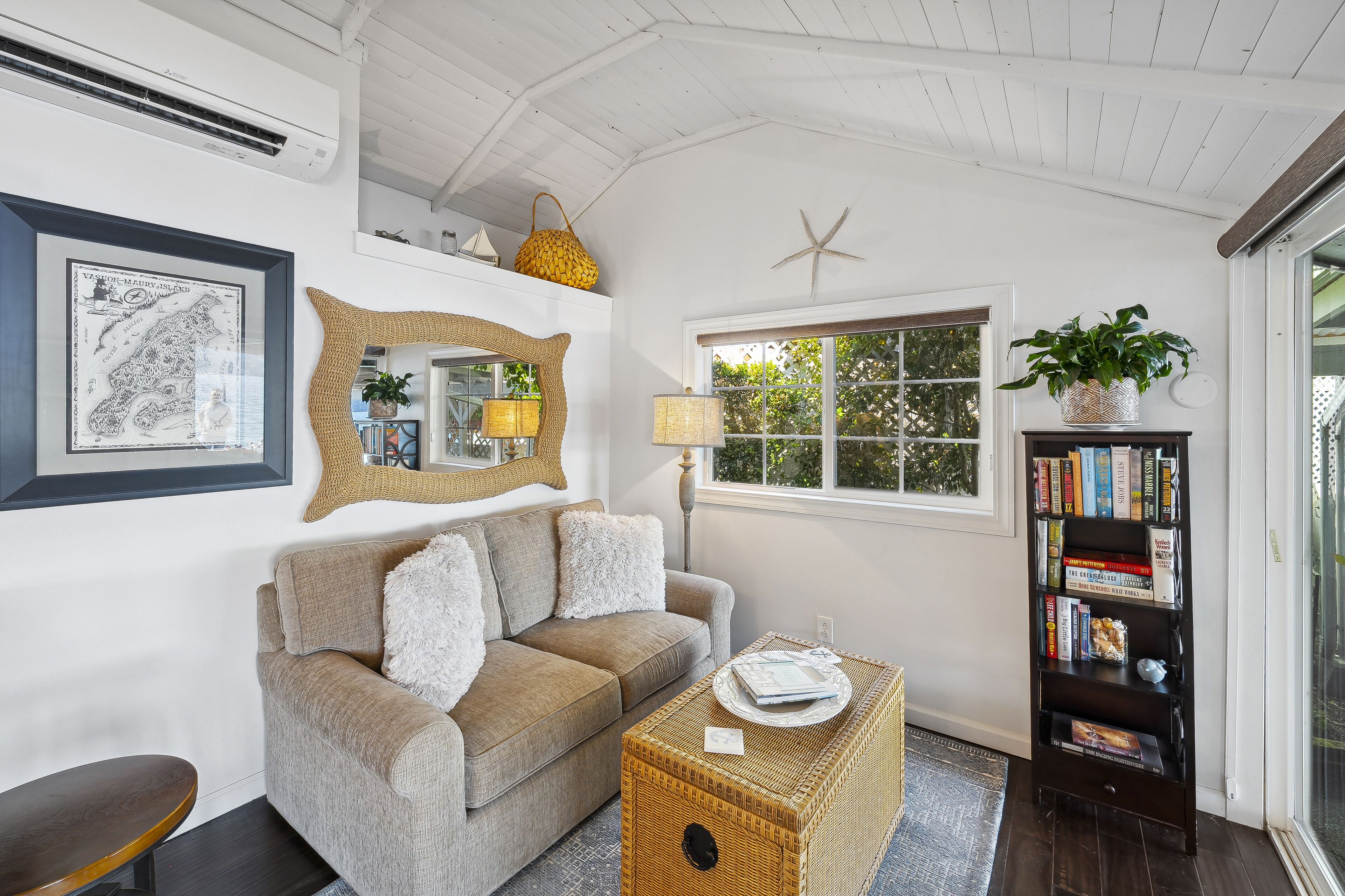 A cozy living room with a sofa, a wicker coffee table, a bookshelf, and a large window with a view of greenery.