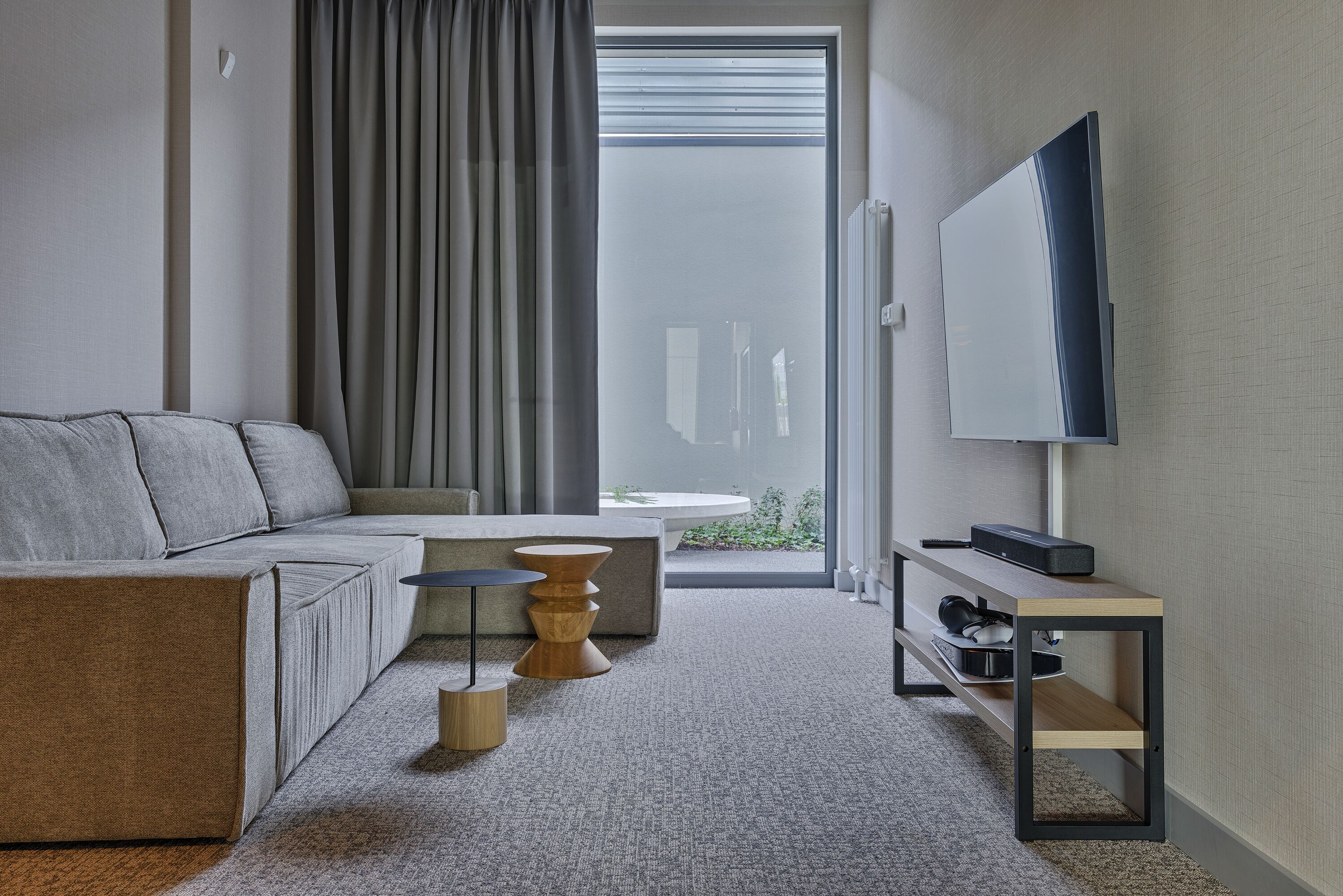 A modern hotel room with a sofa, a small round table, a TV on a stand, and a view of the outdoors through a large window.