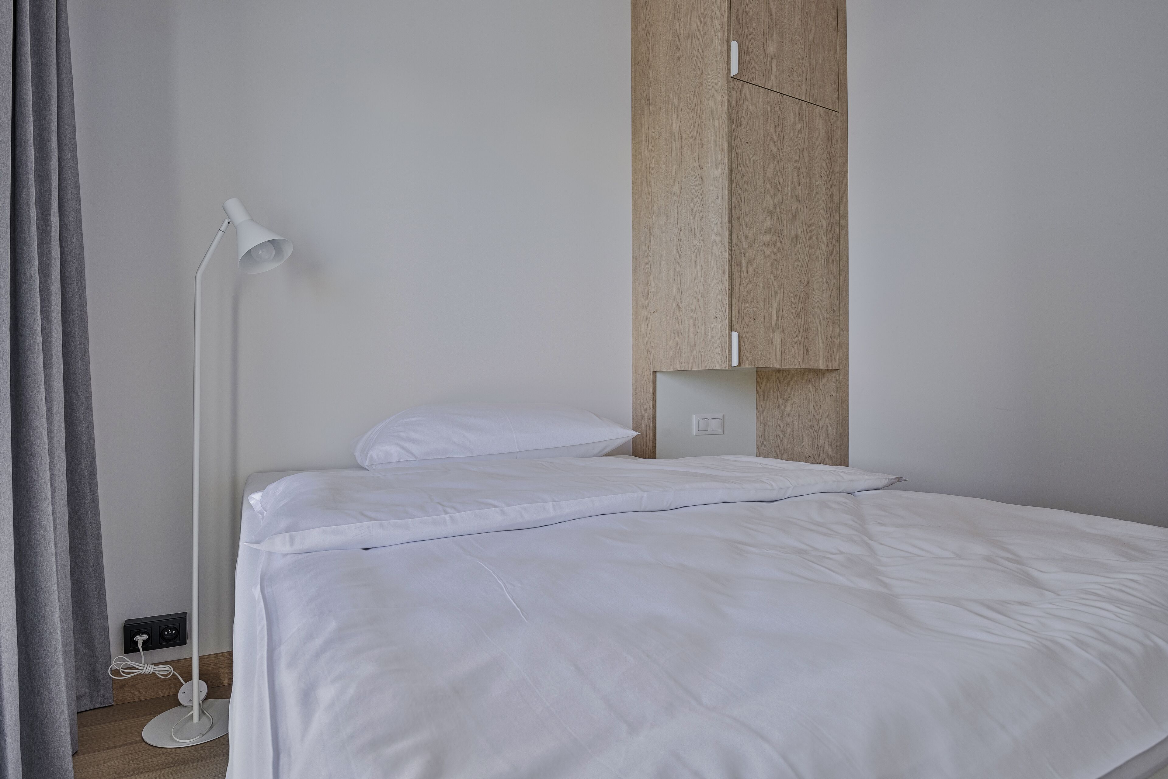 Una cama bien tendida con ropa de cama blanca, una lámpara de pie blanca y un armario de madera en una habitación sencilla y moderna.