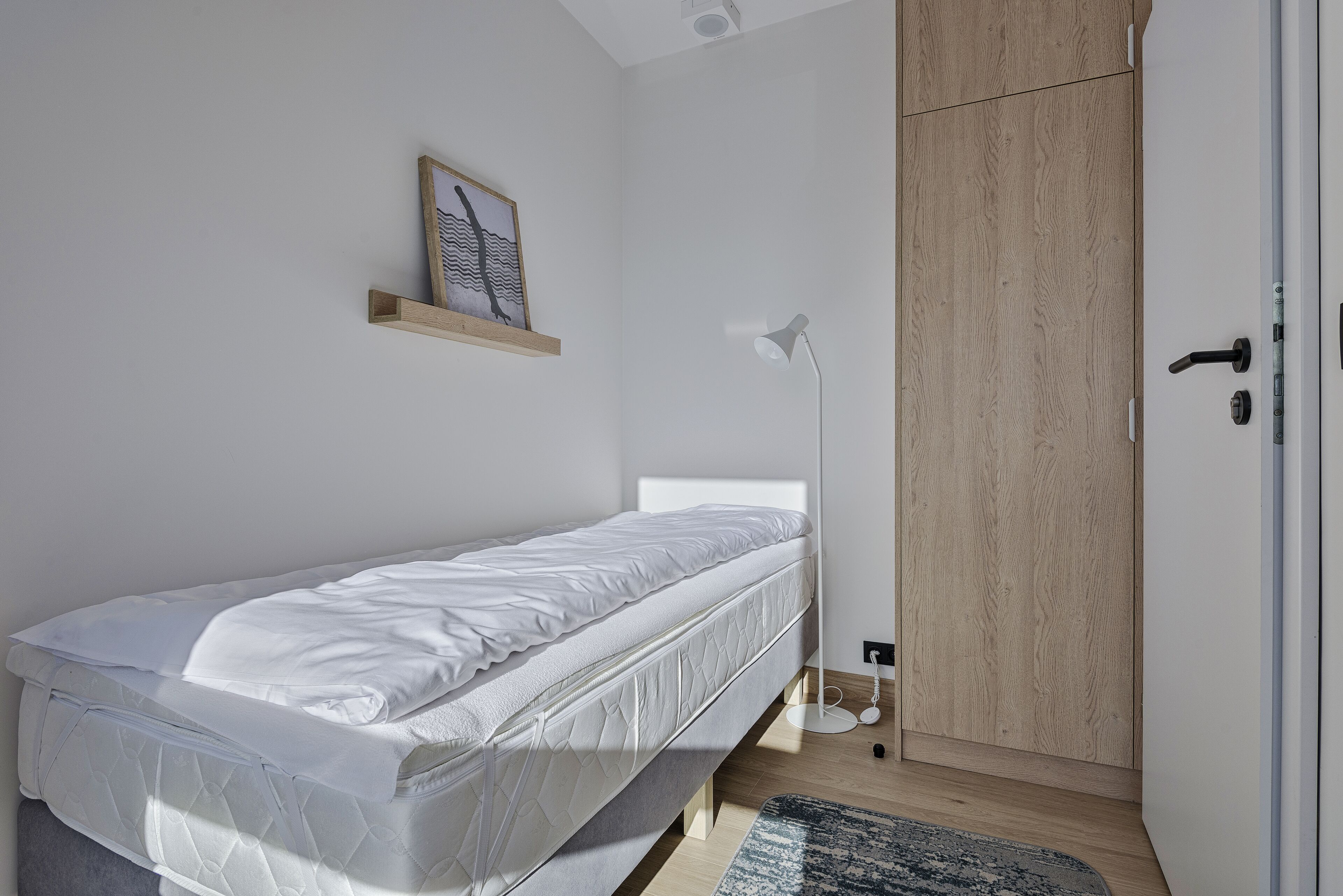 A single bed with a mattress, a bedside lamp, a small shelf with a framed picture, and a wardrobe with a wooden finish.