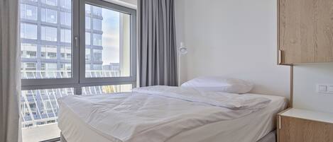 A modern hotel room with a large window, a bed with white linens, a bedside table, and a view of the cityscape.