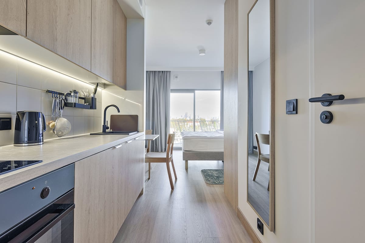 A modern hotel room with a kitchen area, a bed, and a large window offering a city view.