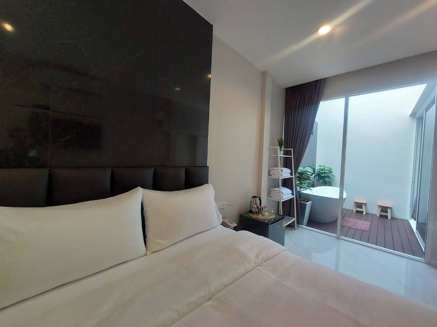 A modern hotel room with a large bed, a nightstand, a glass-enclosed balcony with a view, and a small seating area.